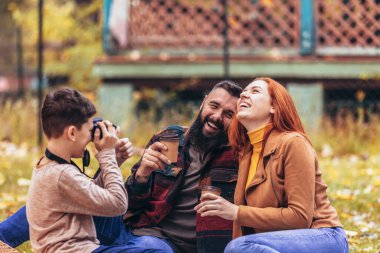 Genç mutlu ebeveynler sonbahar günü parkta oğluyla eğleniyorlar. Çocuk anne ve babasının piknikte fotoğrafını çekiyor. 