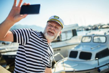 Denizci gömleği ve şapkası giymiş, akıllı telefon kullanarak denizin kenarında duran olgun bir adam.