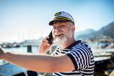 Denizci gömleği ve şapkası giymiş, akıllı telefon kullanarak denizin kenarında duran olgun bir adam.