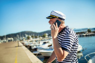 Denizci gömleği ve şapkası giymiş, akıllı telefon kullanarak denizin kenarında duran olgun bir adam.
