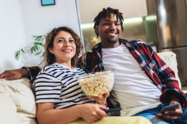 Young happy couple watching movie on TV and eating popcorn while relaxing in the living room.