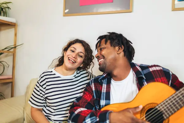 Happy young couple singing a song playing guitar on sofa in living room at home
