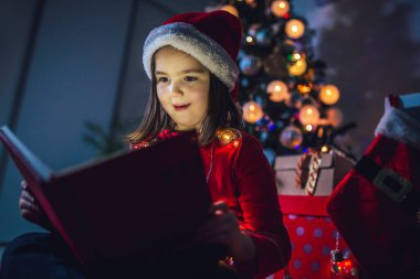 Küçük kız Noel hakkında bir kitap okuyor. Renkli ışıklar, Noel Baba şapkası ve Noel ağacı.