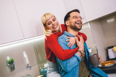 Genç bir adam ve kadın mutfakta dikilip konuşuyorlar. Mutfakta kahve ve meyve suyuyla duran mutlu eş ve koca..