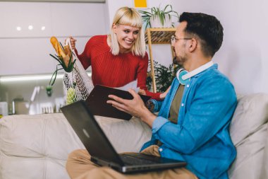 Genç adam kulaklık takıp rahat bir koltukta otururken dizüstü bilgisayarıyla çalışıyor. Karısı marketten yeni geldi, elinde bir torba taze ürünle..