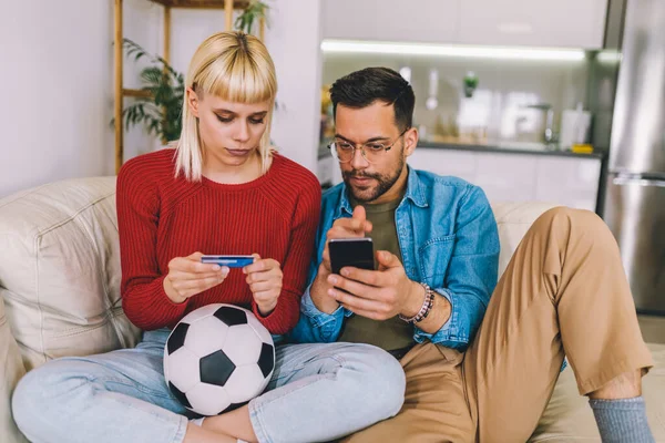 Young couple watching a football game on digital tablet and using credit card for online betting.
