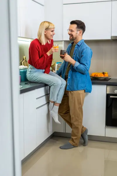 Young man and woman standing in kitchen and talking. Happy wife and husband with cups of coffee and juice standing in the kitchen.