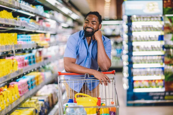 Black Male Buyer Shopping Groceries In Supermarket Taking Product From Shelf Standing With Shop Cart . Looking at the camera.