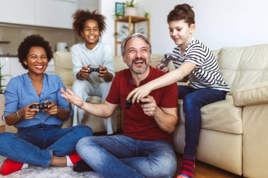 Gülümseyen aile evde birlikte vakit geçiriyorlar. Oturma odasında kanepede oturup video oyunları oynuyorlar..