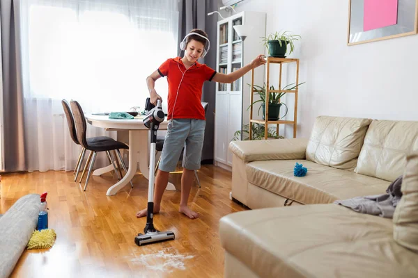 Male child vacuums the dust from the floor with a vacuum cleaner and listening to the music with headphones. Doing his chores.