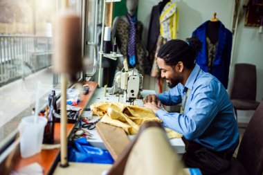 Genç erkek Afrikalı Amerikalı terzi kendi atölyesinde dikiş makinesiyle dikiş dikiyor.