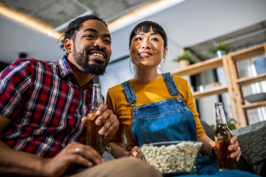 Afro-Amerikalı ve Asyalı çift, kanepede televizyon izleyip, bira içip, patlamış mısır ve pizza yiyorlar..