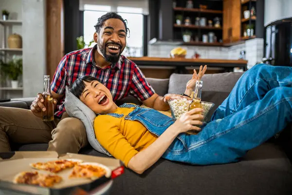 African American and Asian couple, on couch watching tv, drinking beer and eating popcorn and pizza.