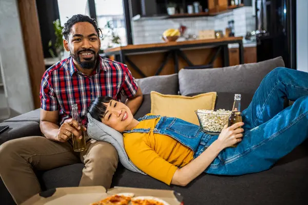 African American and Asian couple, on couch watching tv, drinking beer and eating popcorn and pizza.