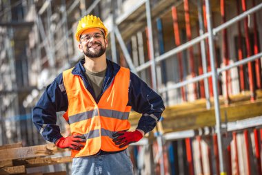 İnşaat alanı yöneticisi çelik yelek ve kask giyip inşaat alanında düşünüyor. Genç mimar inşaat alanını özgüvenle izliyor.