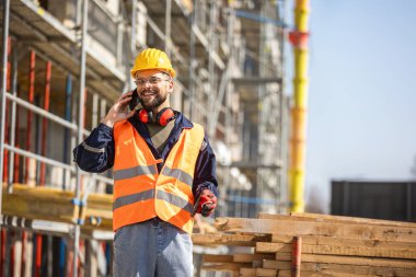 İnşaat alanı yöneticisi çelik yelek ve kask giyip inşaat alanındaki telefonla konuşuyor.. 