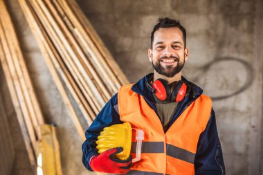 İnşaat alanı yöneticisi çelik yelek ve kask giyip inşaat alanında düşünüyor. Genç mimar inşaat alanını özgüvenle izliyor.
