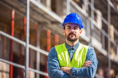 İnşaat alanı yöneticisi çelik yelek ve kask giyip inşaat alanında düşünüyor. Genç mimar inşaat alanını özgüvenle izliyor.