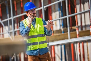 İnşaat alanı yöneticisi çelik yelek ve kask giyip inşaat alanındaki telefonla konuşuyor.. 