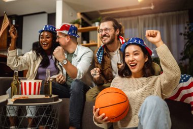 Amerikan bayraklarıyla basketbol maçı izleyen, bira içen ve tezahürat yapan çok ırklı arkadaş grubu..