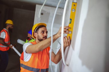 İnşaat işçisi bir inşaat alanında ruh seviyesiyle çalışıyor. Diğer işçiler arka planda konuşuyor..