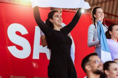 Spor taraftarları stadyumdaki maçta tezahürat yapıyor. Takımlarını desteklemek için mavi ve beyaz renkler giyiyorlar. Bayrak ve eşarplarla kutlama.