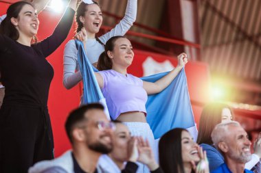 Spor taraftarları stadyumdaki maçta tezahürat yapıyor. Takımlarını desteklemek için mavi ve beyaz renkler giyiyorlar. Bayrak ve eşarplarla kutlama.