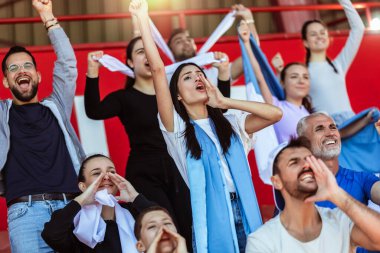 Spor taraftarları stadyumdaki maçta tezahürat yapıyor. Takımlarını desteklemek için mavi ve beyaz renkler giyiyorlar. Bayrak ve eşarplarla kutlama.