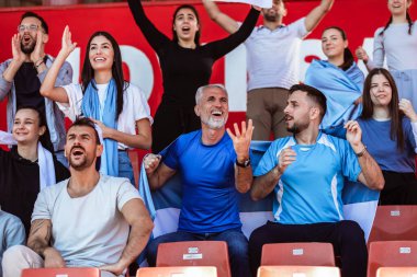 Spor taraftarları stadyumdaki maçta tezahürat yapıyor. Takımlarını desteklemek için mavi ve beyaz renkler giyiyorlar. Bayrak ve eşarplarla kutlama.