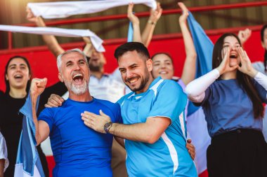 Spor taraftarları stadyumdaki maçta tezahürat yapıyor. Takımlarını desteklemek için mavi ve beyaz renkler giyiyorlar. Bayrak ve eşarplarla kutlama.