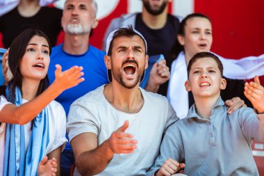 Spor taraftarları stadyumdaki maçta tezahürat yapıyor. Takımlarını desteklemek için mavi ve beyaz renkler giyiyorlar. Bayrak ve eşarplarla kutlama.