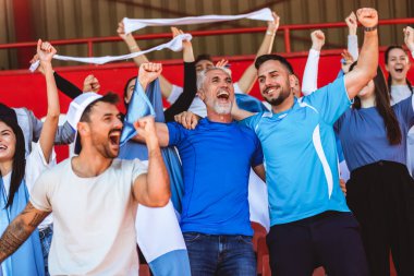 Spor taraftarları stadyumdaki maçta tezahürat yapıyor. Takımlarını desteklemek için mavi ve beyaz renkler giyiyorlar. Bayrak ve eşarplarla kutlama.
