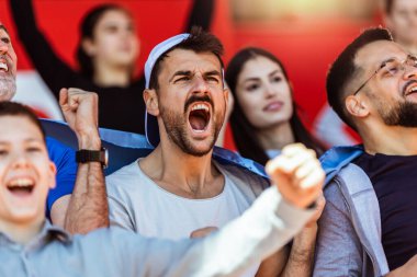 Spor taraftarları stadyumdaki maçta tezahürat yapıyor. Takımlarını desteklemek için mavi ve beyaz renkler giyiyorlar. Bayrak ve eşarplarla kutlama.