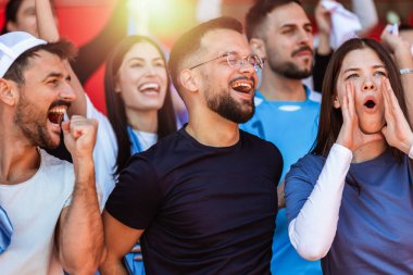 Spor taraftarları stadyumdaki maçta tezahürat yapıyor. Takımlarını desteklemek için mavi ve beyaz renkler giyiyorlar. Bayrak ve eşarplarla kutlama.