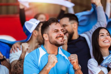 Spor taraftarları stadyumdaki maçta tezahürat yapıyor. Takımlarını desteklemek için mavi ve beyaz renkler giyiyorlar. Bayrak ve eşarplarla kutlama.