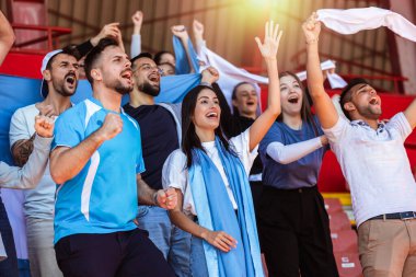 Spor taraftarları stadyumdaki maçta tezahürat yapıyor. Takımlarını desteklemek için mavi ve beyaz renkler giyiyorlar. Bayrak ve eşarplarla kutlama.