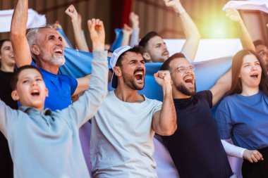Spor taraftarları stadyumdaki maçta tezahürat yapıyor. Takımlarını desteklemek için mavi ve beyaz renkler giyiyorlar. Bayrak ve eşarplarla kutlama.