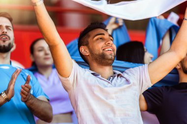 Spor taraftarları stadyumdaki maçta tezahürat yapıyor. Takımlarını desteklemek için mavi ve beyaz renkler giyiyorlar. Bayrak ve eşarplarla kutlama.