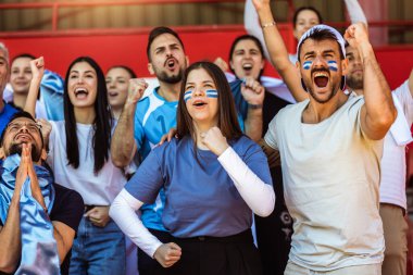 Spor taraftarları stadyumdaki maçta tezahürat yapıyor. Takımlarını desteklemek için mavi ve beyaz renkler giyiyorlar. Bayrak ve eşarplarla kutlama.