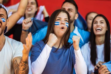 Spor taraftarları stadyumdaki maçta tezahürat yapıyor. Takımlarını desteklemek için mavi ve beyaz renkler giyiyorlar. Bayrak ve eşarplarla kutlama.
