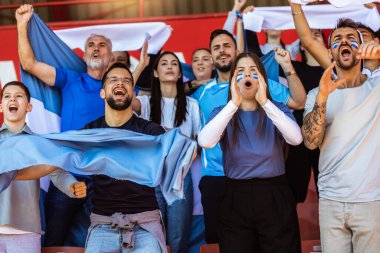 Spor taraftarları stadyumdaki maçta tezahürat yapıyor. Takımlarını desteklemek için mavi ve beyaz renkler giyiyorlar. Bayrak ve eşarplarla kutlama.