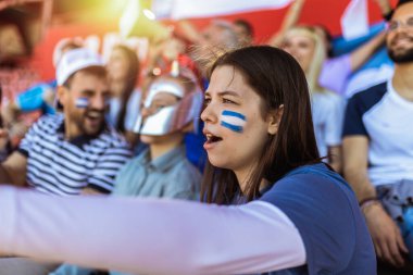 Spor taraftarları stadyumdaki maçta tezahürat yapıyor. Takımlarını desteklemek için mavi ve beyaz renkler giyiyorlar. Bayrak ve eşarplarla kutlama.
