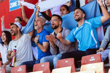 Spor taraftarları stadyumdaki maçta tezahürat yapıyor. Takımlarını desteklemek için mavi ve beyaz renkler giyiyorlar. Bayrak ve eşarplarla kutlama.