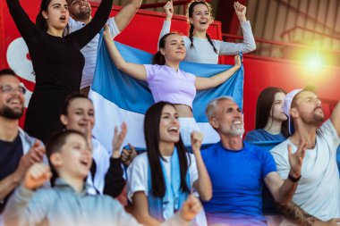 Spor taraftarları stadyumdaki maçta tezahürat yapıyor. Takımlarını desteklemek için mavi ve beyaz renkler giyiyorlar. Bayrak ve eşarplarla kutlama.