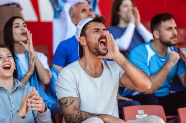 Spor taraftarları stadyumdaki maçta tezahürat yapıyor. Takımlarını desteklemek için mavi ve beyaz renkler giyiyorlar. Bayrak ve eşarplarla kutlama.