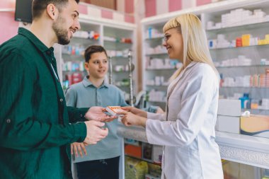 Neşeli genç sarışın eczacı kadın vitamin ve ilaç veriyor baba ve oğluna.