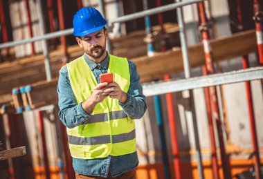 İnşaat alanı yöneticisi çelik yelek ve kask giyip inşaat alanında telefonunu kullanıyor..