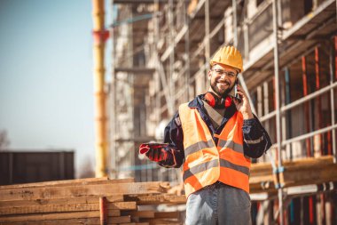 İnşaat alanı yöneticisi çelik yelek ve kask giyip inşaat alanındaki telefonla konuşuyor..