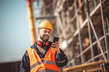 İnşaat alanı yöneticisi çelik yelek ve kask giyip inşaat alanında telefonunu kullanıyor..