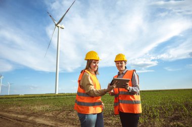 İki kadın mühendis rüzgar türbini çiftliğinde çalışıyor ve raporlara bakıyor. Korunmak için floresan yelek ve kask giyiyorlar..